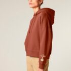 Sweat bio femme avec capuche de couleur terracotta avec le dessin de la marque sur la manche représentant un homme qui grimpe sur un arbre au dessus de la planète. La couleur de sérigraphie est beige.