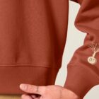 Sweat bio femme de couleur terracotta avec le dessin de la marque sur la manche représentant un homme qui grimpe sur un arbre au dessus de la planète. La couleur de sérigraphie est beige.