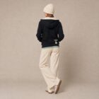 Photo de présentation du sweat à capuche zippé unisexe Polaire, en coton biologique, coloris noir, porté par un modèle femme. Collection Ardecho 07. Vue de dos avec le dessin de la marque en bas à droite, représentant un arbre sur la planète et un homme qui grimpe à l'arbre, la sérigraphie est de couleur beige.
