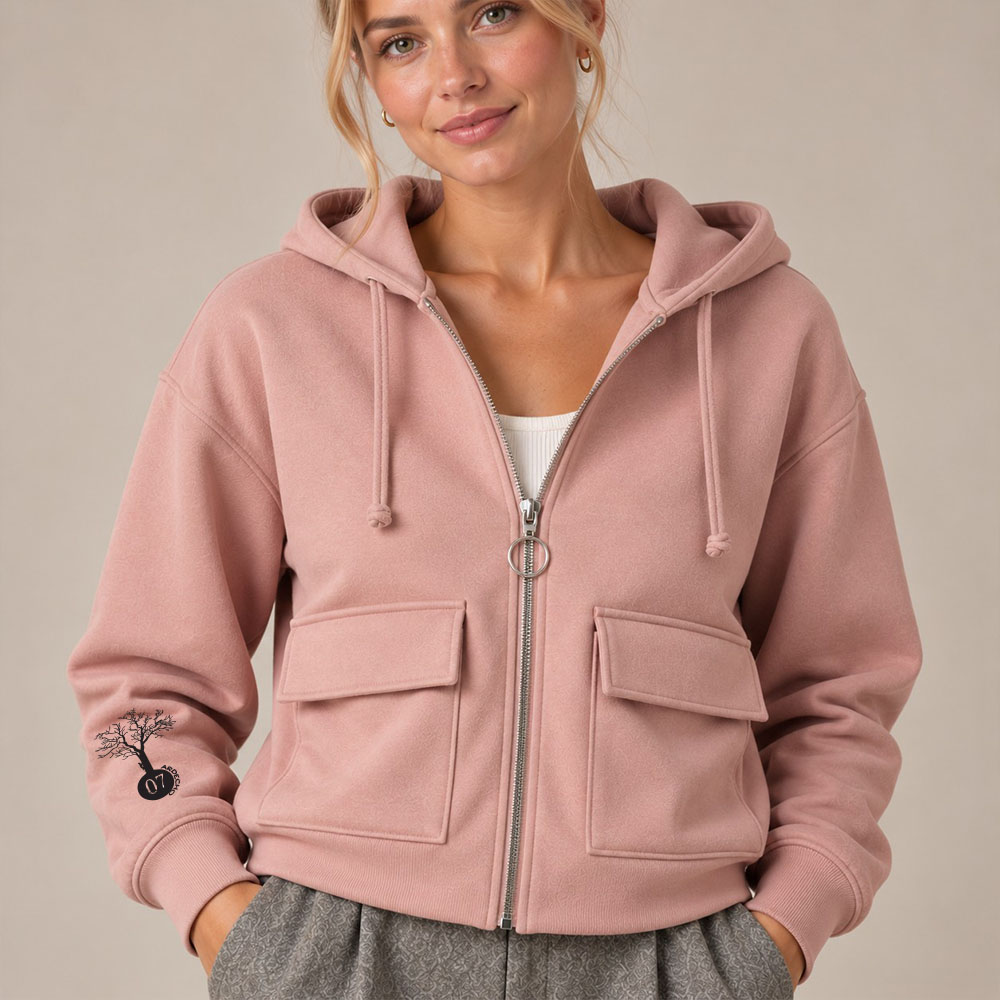 Sweat coton bio femme avec une capuche couleur rosé et une fermeture éclaire. Vue de face. Dessin de la marque sur la manche de couleur noir, représentant un arbre sur la planète et une personne qui grimpe à l'arbre. Zoom sur la veste.