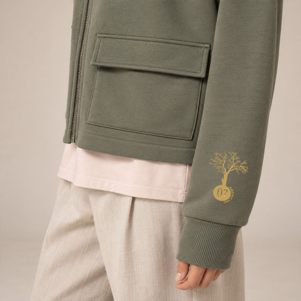 Sweat coton bio femme avec une capuche couleur kaki et une fermeture éclaire. Vue de coté. Dessin de la marque sur la manche de couleur doré, représentant un arbre sur la planète et une personne qui grimpe à l'arbre. Zoom sur la veste.