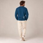 Sweat homme en coton biologique de couleur bleu denim avec le dessin de la marque, en bas dans le dos représentant un arbre sur une planète et un homme qui grimpe à l'arbre. la sérigraphie est beige.